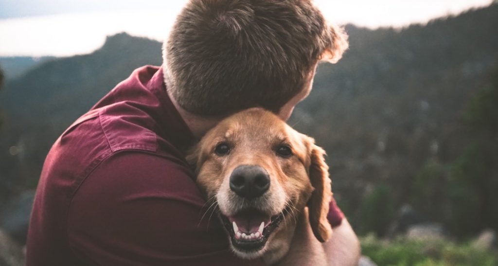A man wearing a red shirt is hugging a dog outside that is smiling