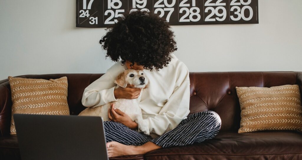 A woman sitting on a couch is kissing a dog on the top of their head