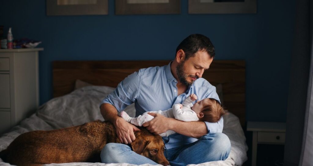A man is holding a baby with a dog in bed