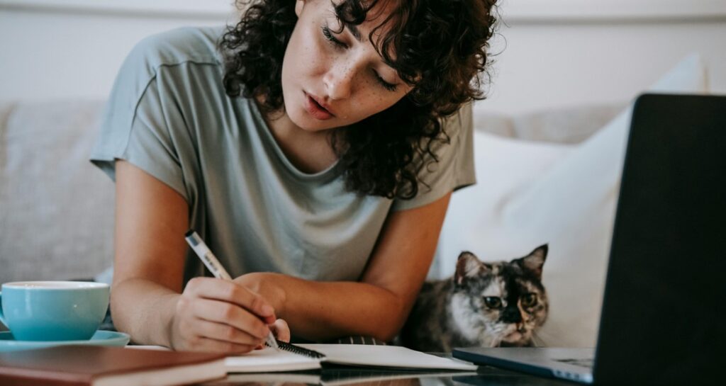 A woman is writing in a notebook while sitting on a couch with their cat